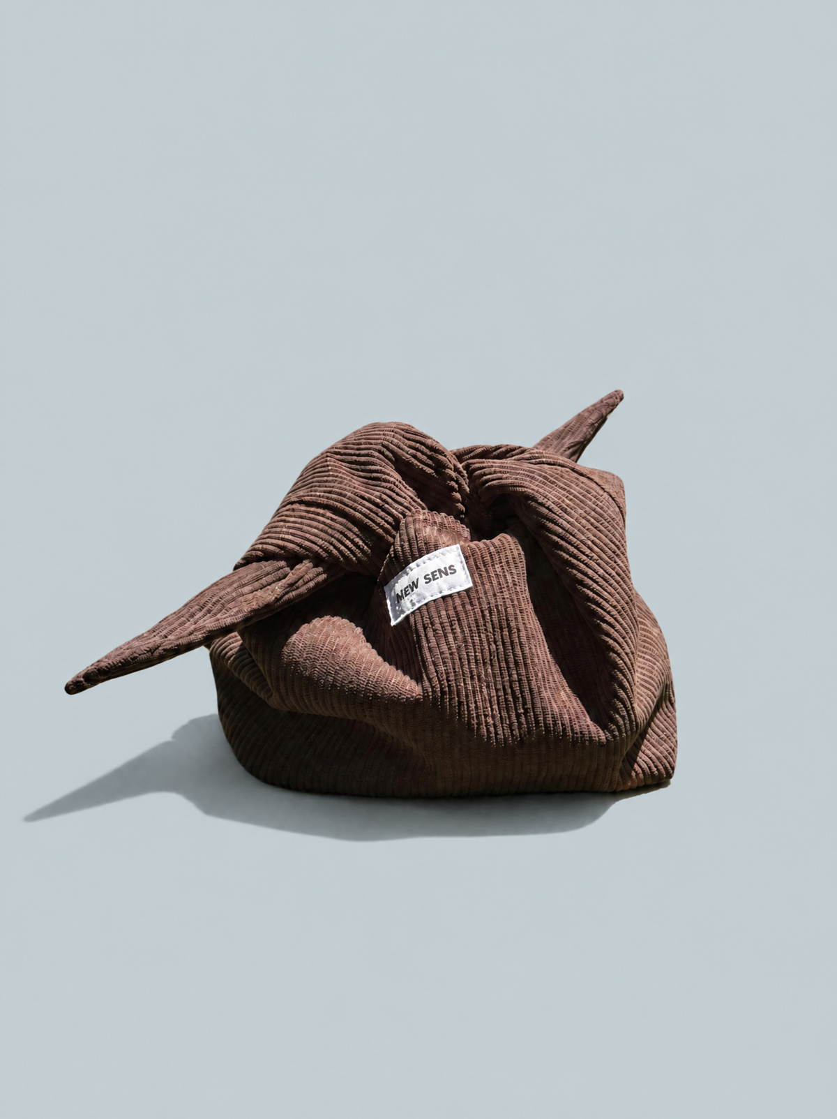A brown corduroy gift bag tied in a knot, featuring a white label that reads 'NEW SENS', set against a solid light gray background.